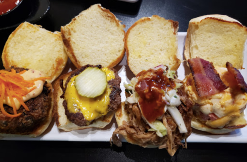 A platter with four mini sandwiches: a burger with sauce, a cheeseburger, pulled pork with slaw, and a bacon-topped sandwich.