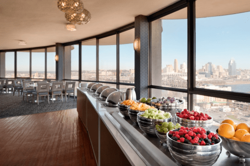 A buffet of fresh fruits in silver bowls, with a panoramic city view through large windows.