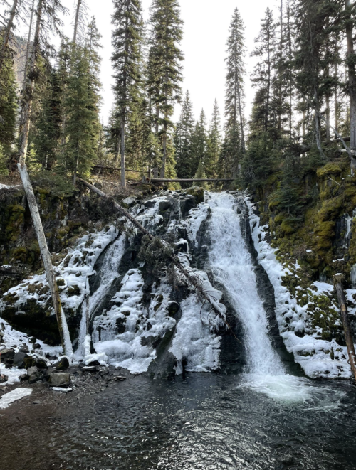 A serene waterfall cascades over rocks, surrounded by evergreen trees and patches of snow.