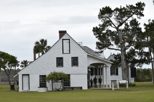 A white historic house with a porch, surrounded by trees and grass, under a cloudy sky.