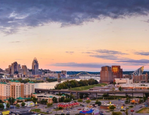 Panoramic view of a city skyline at sunset, with buildings, a river, and a colorful sky.