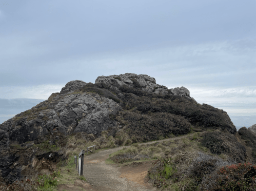 A rocky outcrop surrounded by grassy terrain under a cloudy sky, with a winding path leading through the landscape.
