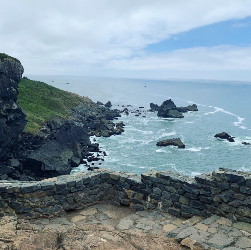 A rocky coastline with waves crashing against the shore, under a cloudy sky. Green hills rise in the background.