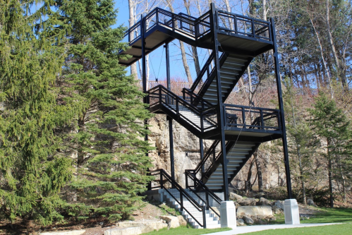 A modern black metal staircase with multiple levels, surrounded by trees and rocky terrain.