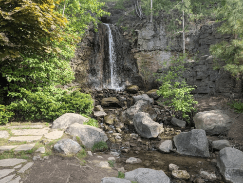 A serene waterfall cascades over rocks, surrounded by lush greenery and boulders in a peaceful natural setting.