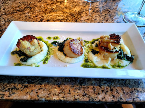 Three seared scallops on a bed of creamy mashed potatoes, garnished with greens and a drizzle of sauce, on a white plate.