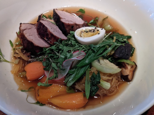A bowl of noodles topped with sliced meat, vegetables, and a soft-boiled egg in a savory broth.