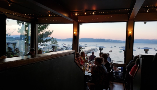 A cozy restaurant with large windows overlooking a lake at sunset, with diners enjoying their meals inside.
