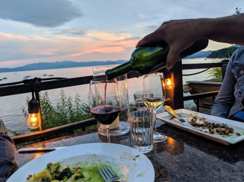 A hand pours red wine into a glass at a scenic waterfront dining table during sunset. Plates of food are visible.