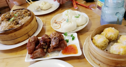 A table spread with dim sum, including dumplings, buns, fried chicken, and noodles, garnished with herbs.