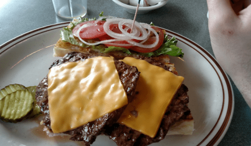 A plate with a sandwich featuring two beef patties, melted cheese, lettuce, tomato, and onion, alongside pickles.