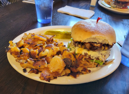 A plate with a burger topped with lettuce and a cherry, served with crispy hash browns and a pickle on the side.