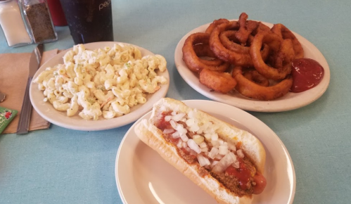 A plate with a chili dog topped with onions, a side of macaroni salad, and a serving of crispy onion rings with ketchup.