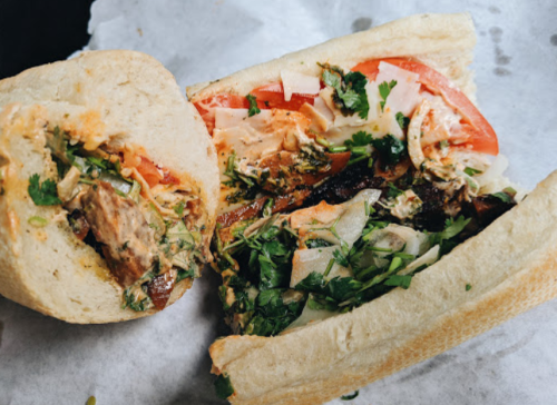 A close-up of a sandwich filled with grilled meat, tomatoes, onions, and cilantro, served in a soft bread roll.
