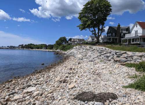 A rocky shoreline with houses and trees in the background under a blue sky with scattered clouds.