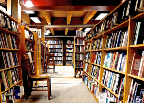 A cozy bookstore aisle lined with bookshelves, featuring a wooden chair and warm lighting.