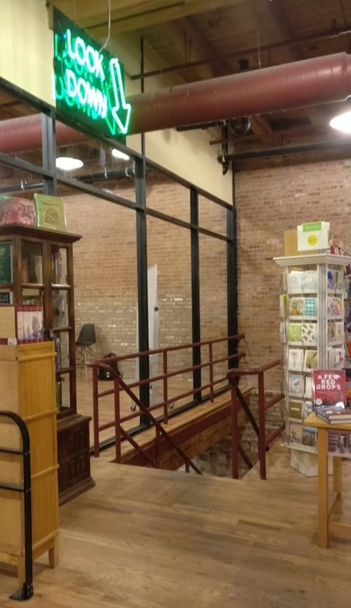 Interior of a bookstore with a "LOOK DOWN" neon sign, wooden flooring, and shelves displaying books and magazines.