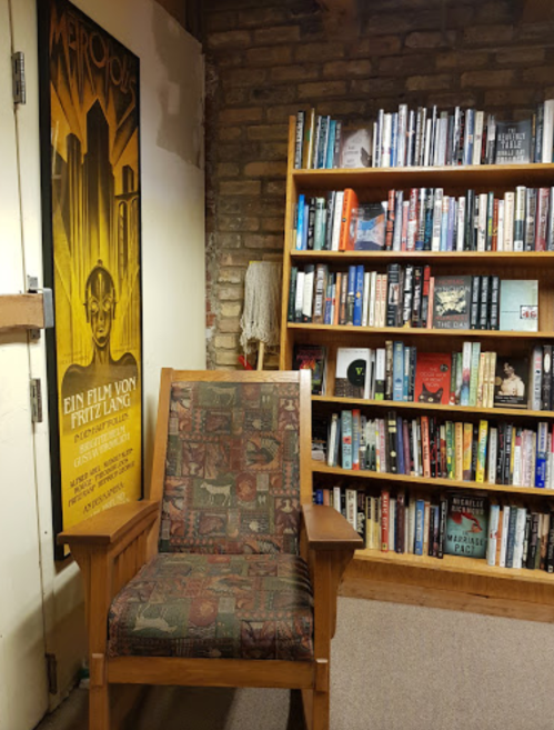 A cozy reading nook featuring a patterned chair next to a bookshelf filled with various books and a vintage movie poster.