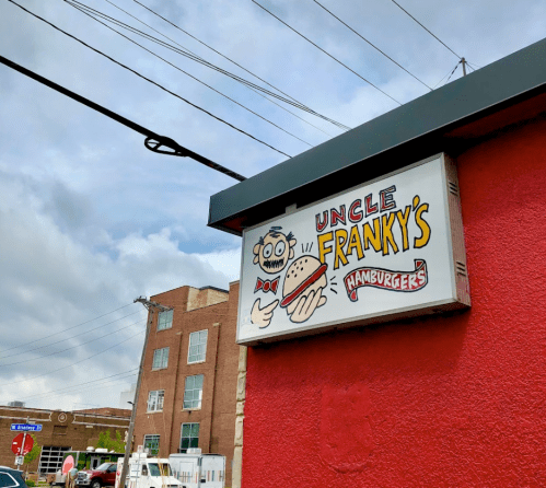 Sign for "Uncle Franky's Hamburgers" featuring a cartoon character holding a burger, against a red wall and cloudy sky.