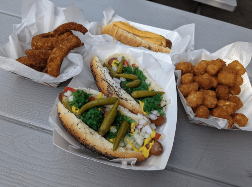A variety of hot dogs with toppings, crispy snacks, and tater tots served in paper trays on a table.
