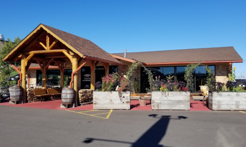 A rustic building with a wooden awning, surrounded by flower planters and barrels, set against a clear blue sky.