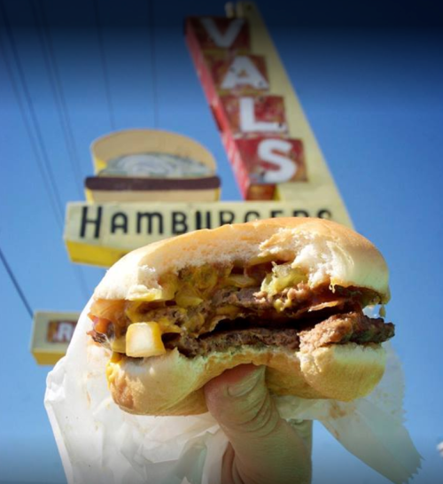 A hand holds a juicy hamburger in front of a retro sign for Val's Hamburgers against a clear blue sky.