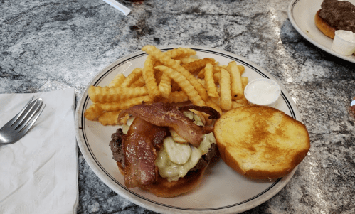 A plate with a bacon cheeseburger and crinkle-cut fries, accompanied by a small cup of dipping sauce.