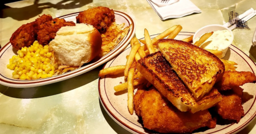 A plate of fried chicken, macaroni and cheese, and bread rolls next to a plate of grilled fish and fries with dipping sauce.