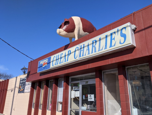 A red and white building with a sign reading "Cheap Charlie's" and a large pig statue on the roof against a blue sky.