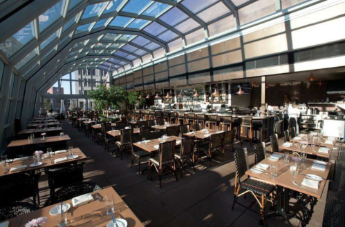 Bright, modern restaurant interior with glass ceiling, wooden tables, and a bar in the background.