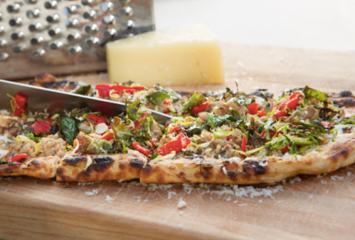 A freshly baked flatbread pizza topped with greens, red peppers, and cheese, with a grater and cheese block in the background.