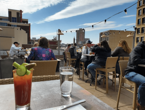 A rooftop bar scene with patrons enjoying drinks, a city skyline in the background, and a clear blue sky.
