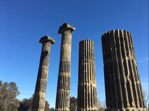 Four ancient stone columns stand against a clear blue sky, surrounded by trees in the background.