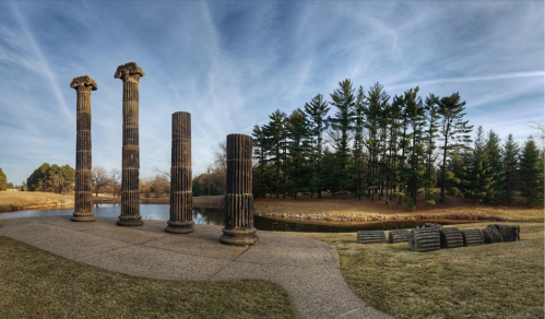 Four ancient columns stand near a pond, surrounded by trees and a grassy area, under a blue sky with wispy clouds.