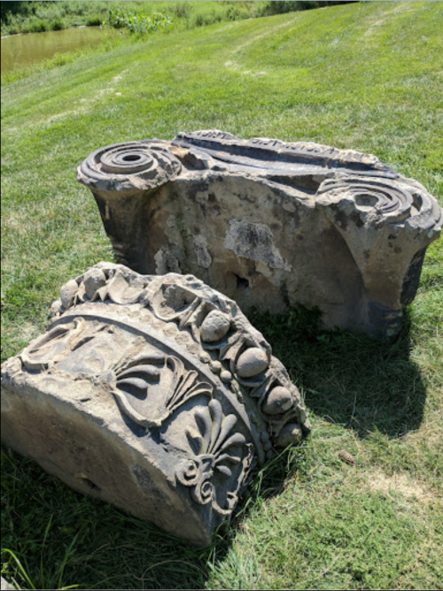 Two weathered stone fragments with intricate carvings, resting on grass near a body of water.