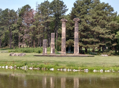 Four ancient columns stand in a grassy area by a calm pond, surrounded by trees and greenery.