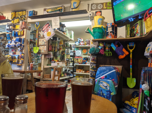 Two glasses of dark beer on a wooden table, surrounded by shelves filled with tools and toys, with a TV showing a soccer game.