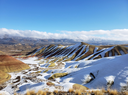 Colorful, layered hills covered in snow under a clear blue sky, with distant mountains in the background.
