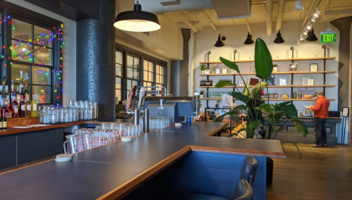 A modern bar with a dark countertop, shelves of books, and colorful lights in the windows. A person is seen in the background.