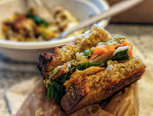 A close-up of a sandwich with greens and orange filling, resting on brown paper, with a bowl of food in the background.