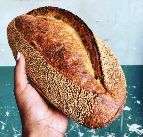 A hand holding a golden-brown loaf of bread covered in sesame seeds against a textured blue and green background.