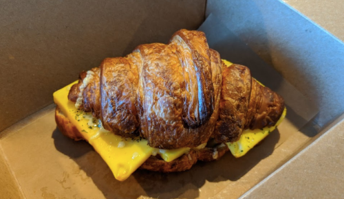A golden-brown croissant sandwich with melted cheese inside, placed in a brown takeout box.