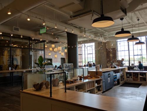 A modern café interior with large windows, wooden counters, and hanging lights, featuring customers enjoying their time.