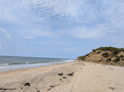 A sandy beach with gentle waves, cliffs in the background, and a cloudy blue sky.