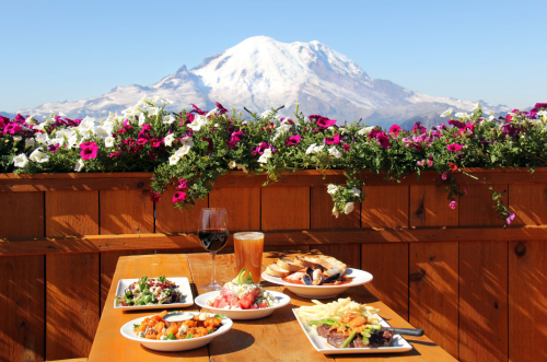 A scenic outdoor dining setup with various dishes and drinks, overlooking a majestic snow-capped mountain and colorful flowers.