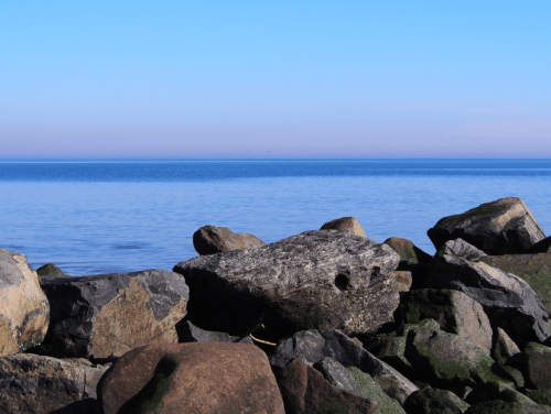A serene seascape featuring smooth rocks in the foreground and calm blue waters under a clear sky.