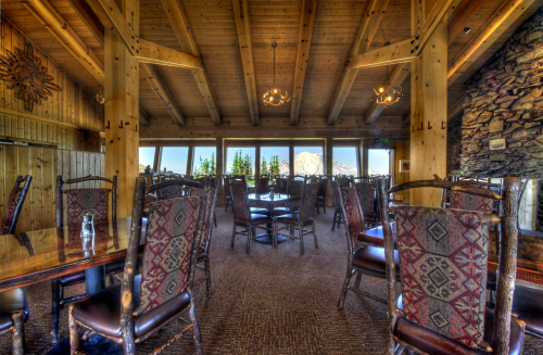 Cozy restaurant interior with wooden beams, stone fireplace, and tables overlooking a mountain view through large windows.