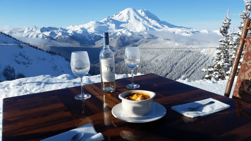 A scenic mountain view with a table set for two, featuring a bowl of soup and wine glasses against a snowy backdrop.