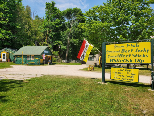A rustic roadside stand offers fresh fish, beef jerky, and local snacks, surrounded by trees and a welcoming atmosphere.