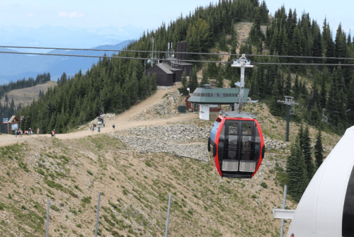 A red gondola glides over a mountainous landscape with trees and a building in the background.